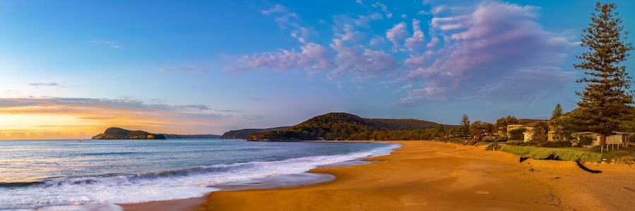 A beach with a sandy shore, ocean waves, a mountain, and houses along the coastline.