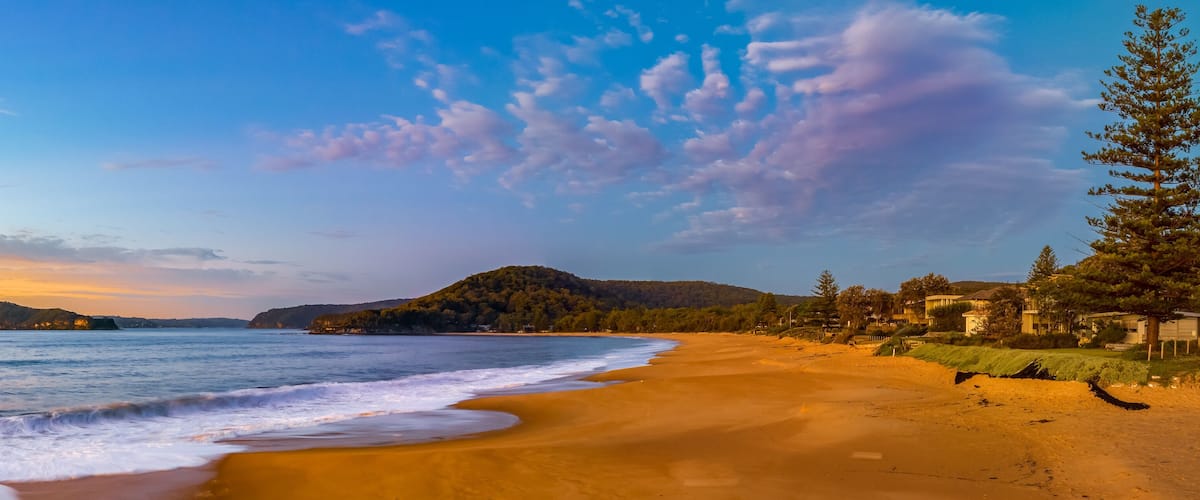 A beach with a sandy shore, ocean waves, a mountain, and houses along the coastline.