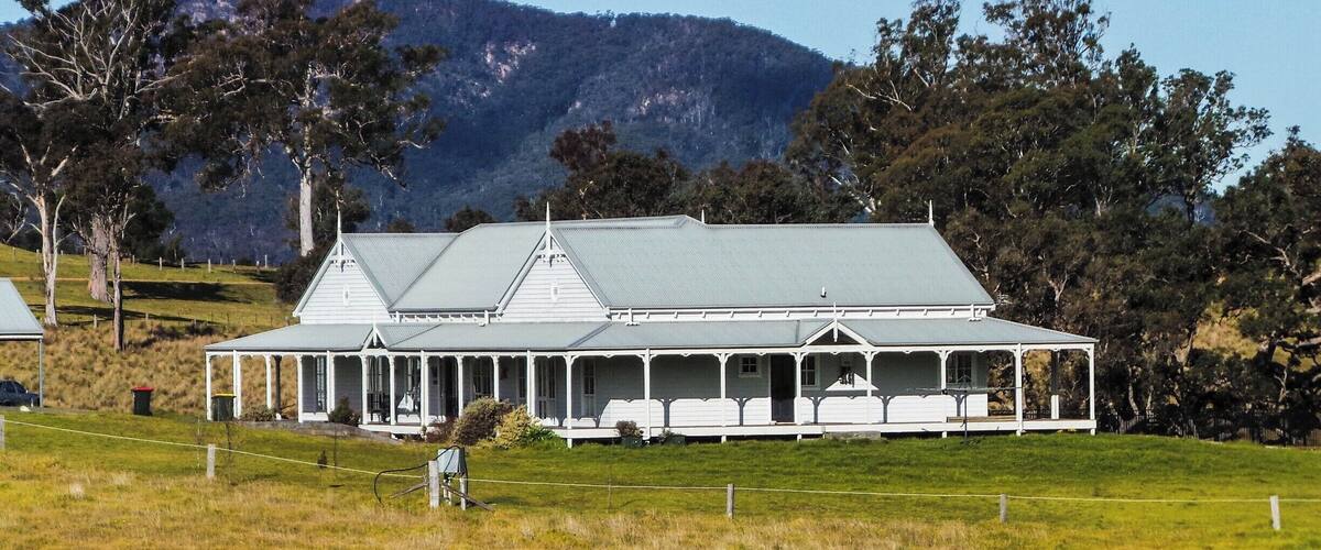A large white house with a green roof, surrounded by a grassy field and trees, with a mountain in the background.