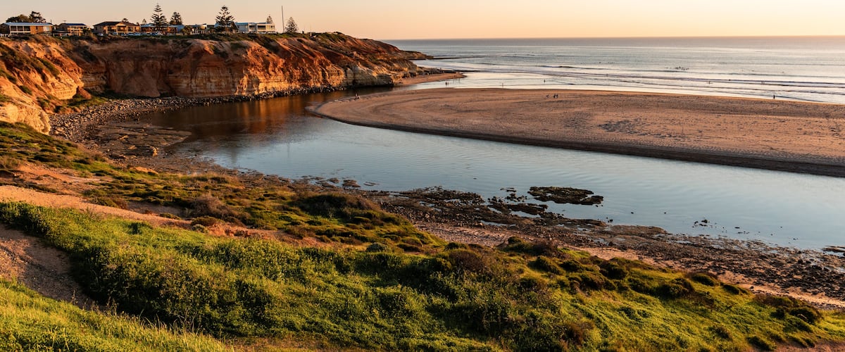 A coastal landscape with a sandy beach, a calm bay, and a cliff with buildings on top.