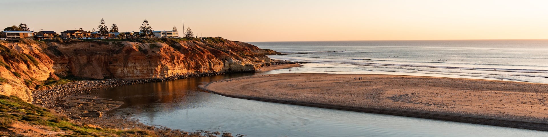 A coastal landscape with a sandy beach, a calm bay, and a cliff with buildings on top.