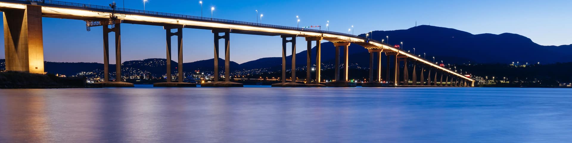 A long bridge spanning a calm body of water with rocky outcrops in the foreground.