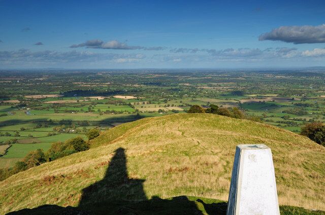 A grassy hill with a stone marker and a view of a valley with fields and distant buildings.