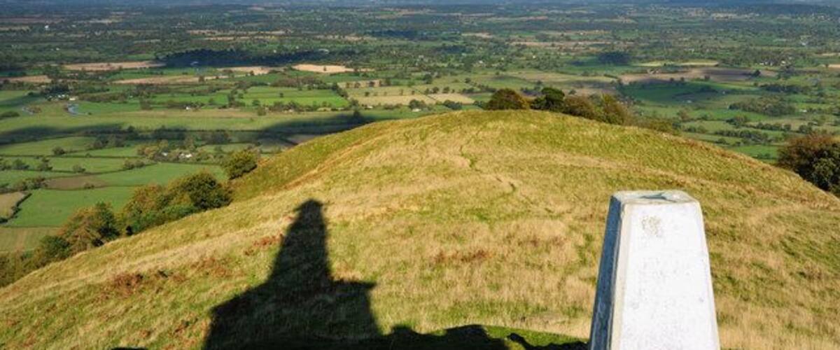 A grassy hill with a stone marker and a view of a valley with fields and distant buildings.