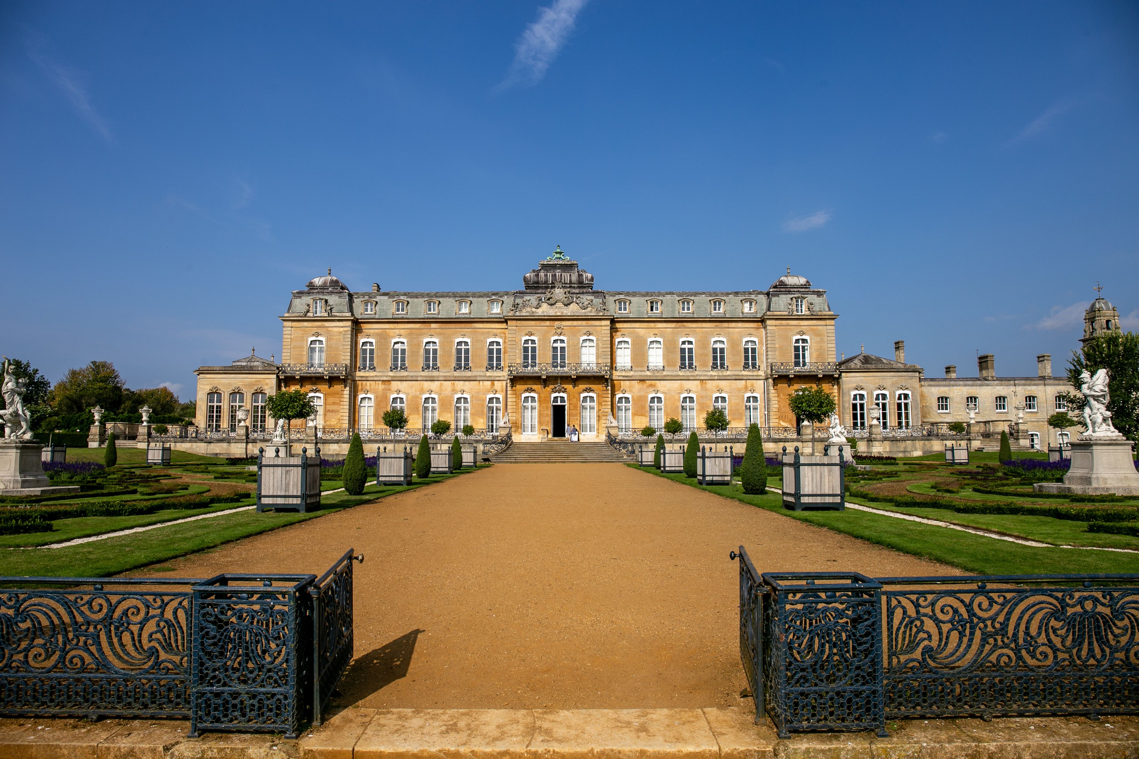 A large, historic building with a symmetrical facade, a central entrance, and a formal garden with statues and trimmed hedges.