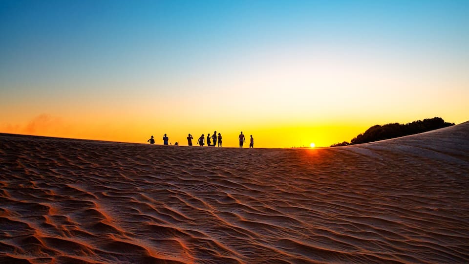 A desert landscape with a group of people standing on a sand dune during sunset.