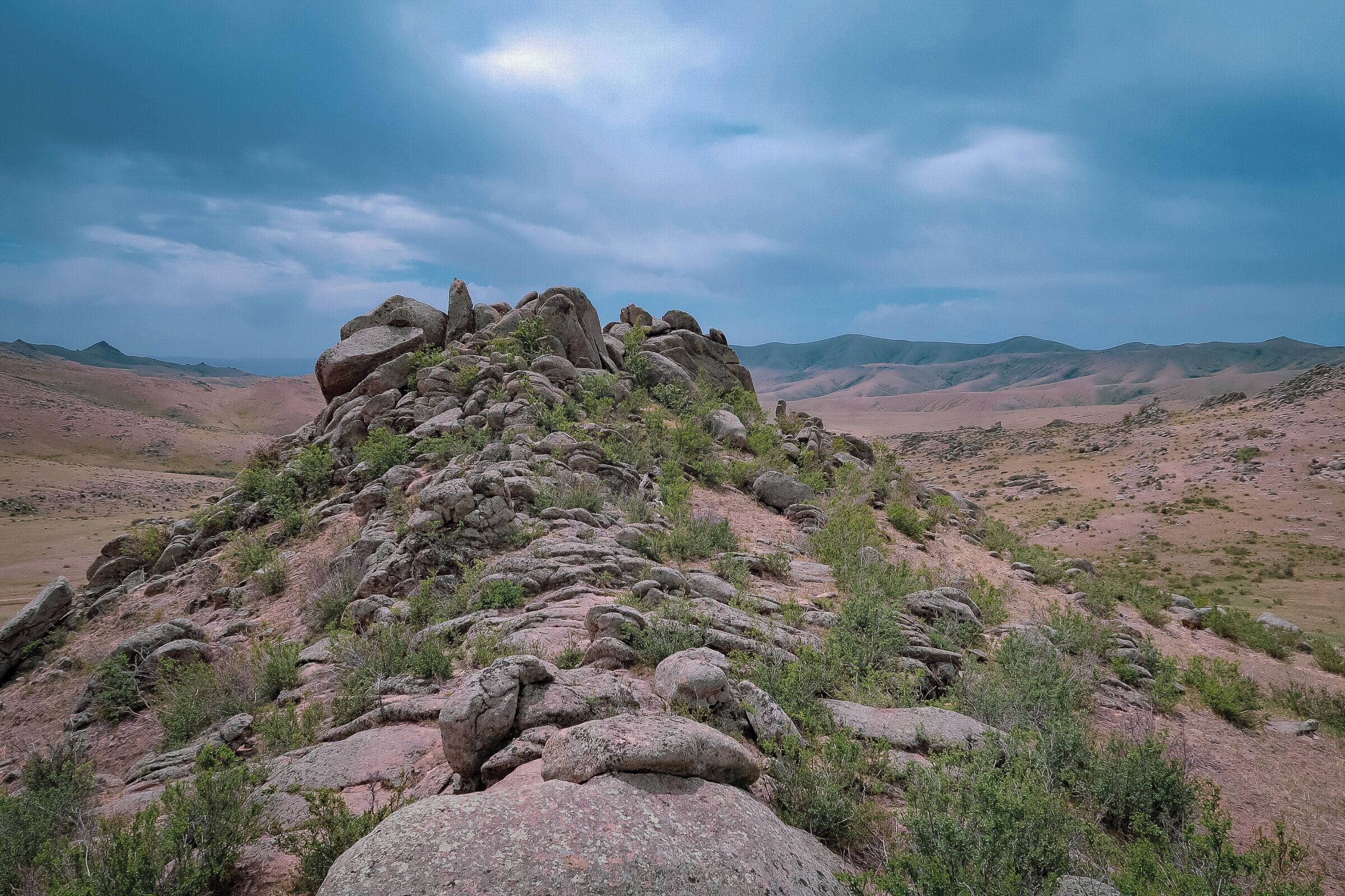 A rocky hill with sparse vegetation and distant mountains under a cloudy sky.