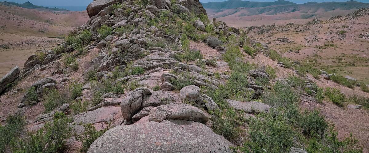 A rocky hill with sparse vegetation and distant mountains under a cloudy sky.