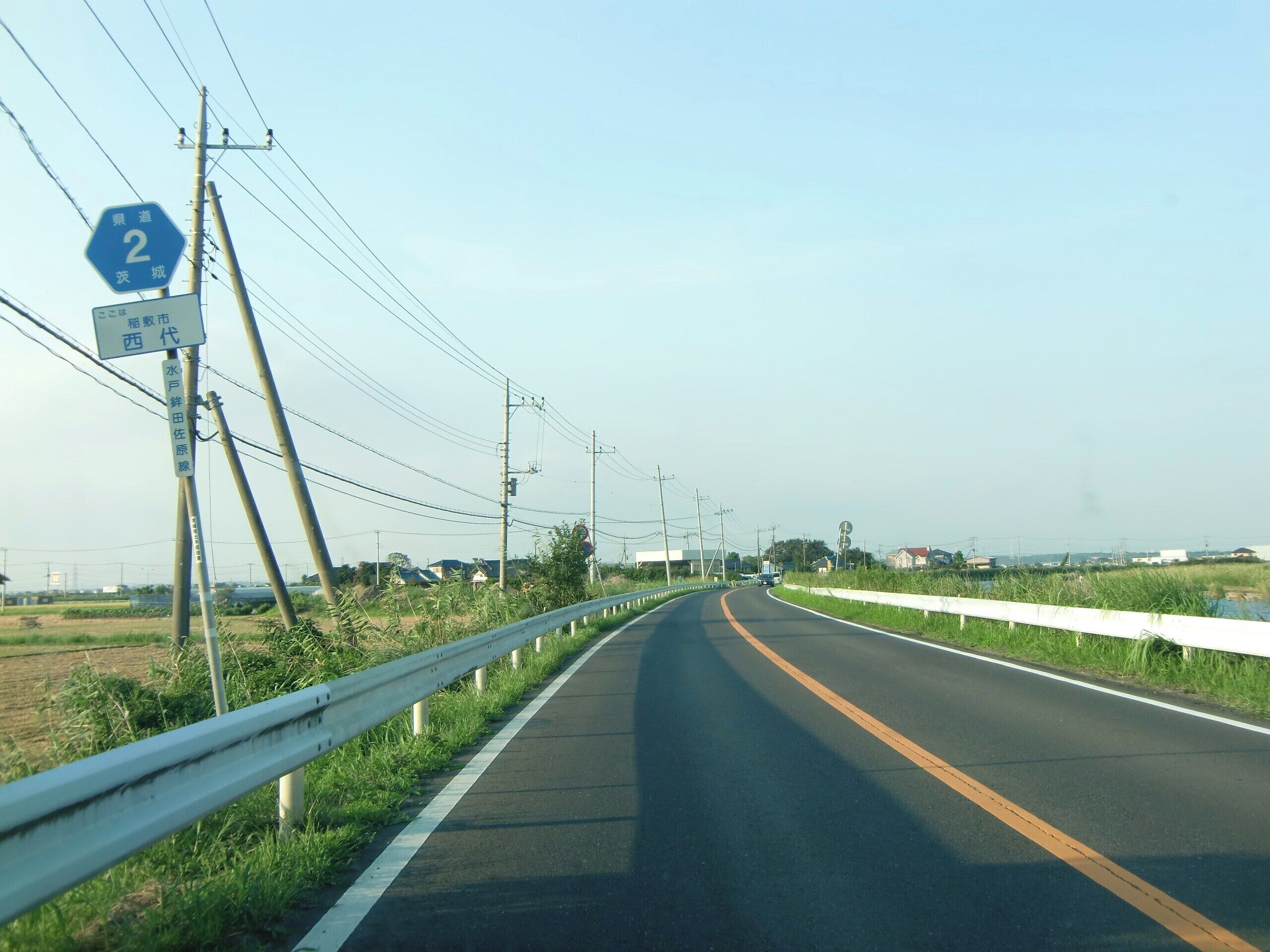 A rural road with a guardrail, utility poles, and a blue sign indicating a route number.