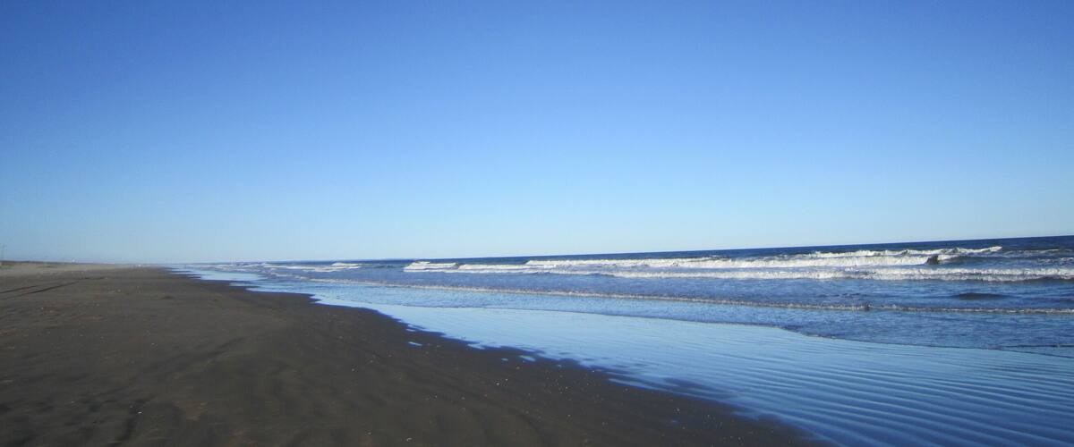 A beach with dark sand and gentle waves under a clear blue sky.