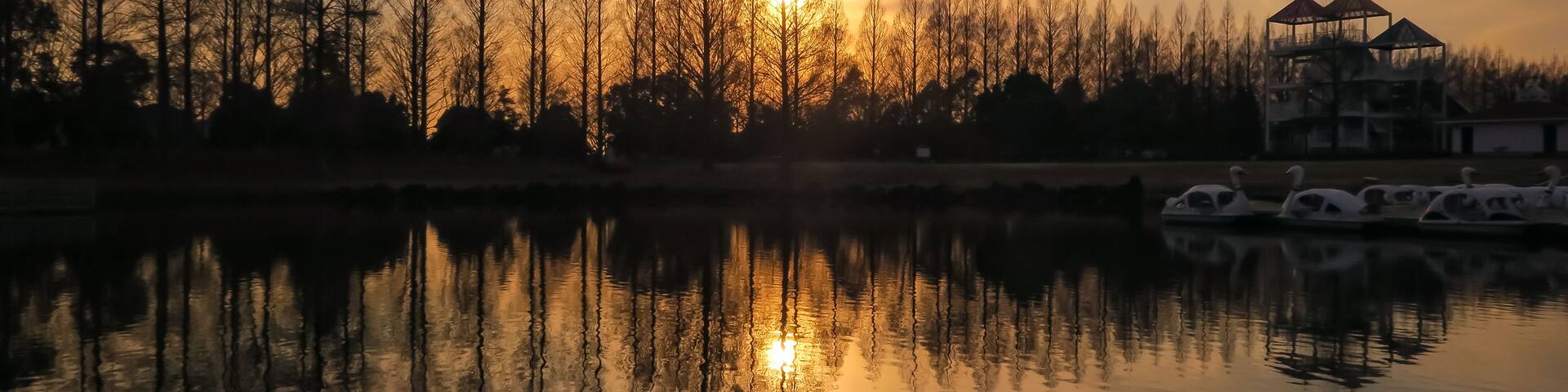 A sunset over a calm lake with reflections of trees and a gazebo.