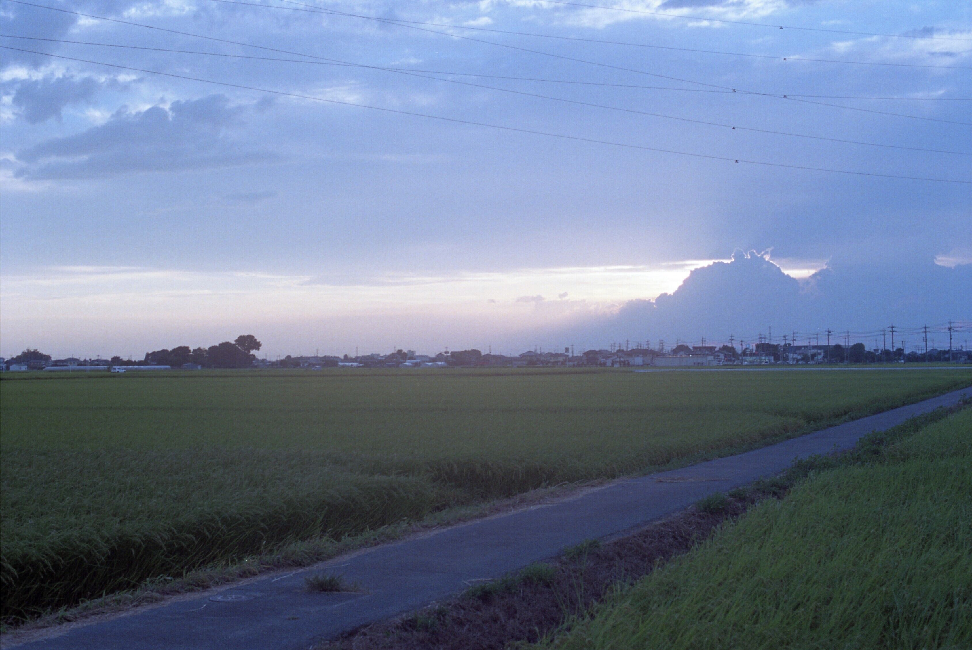 A rural landscape with a road, fields, and distant buildings under a cloudy sky.