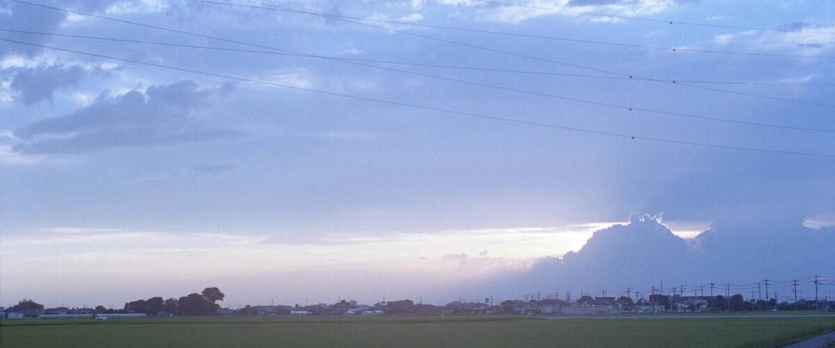 A rural landscape with a road, fields, and distant buildings under a cloudy sky.