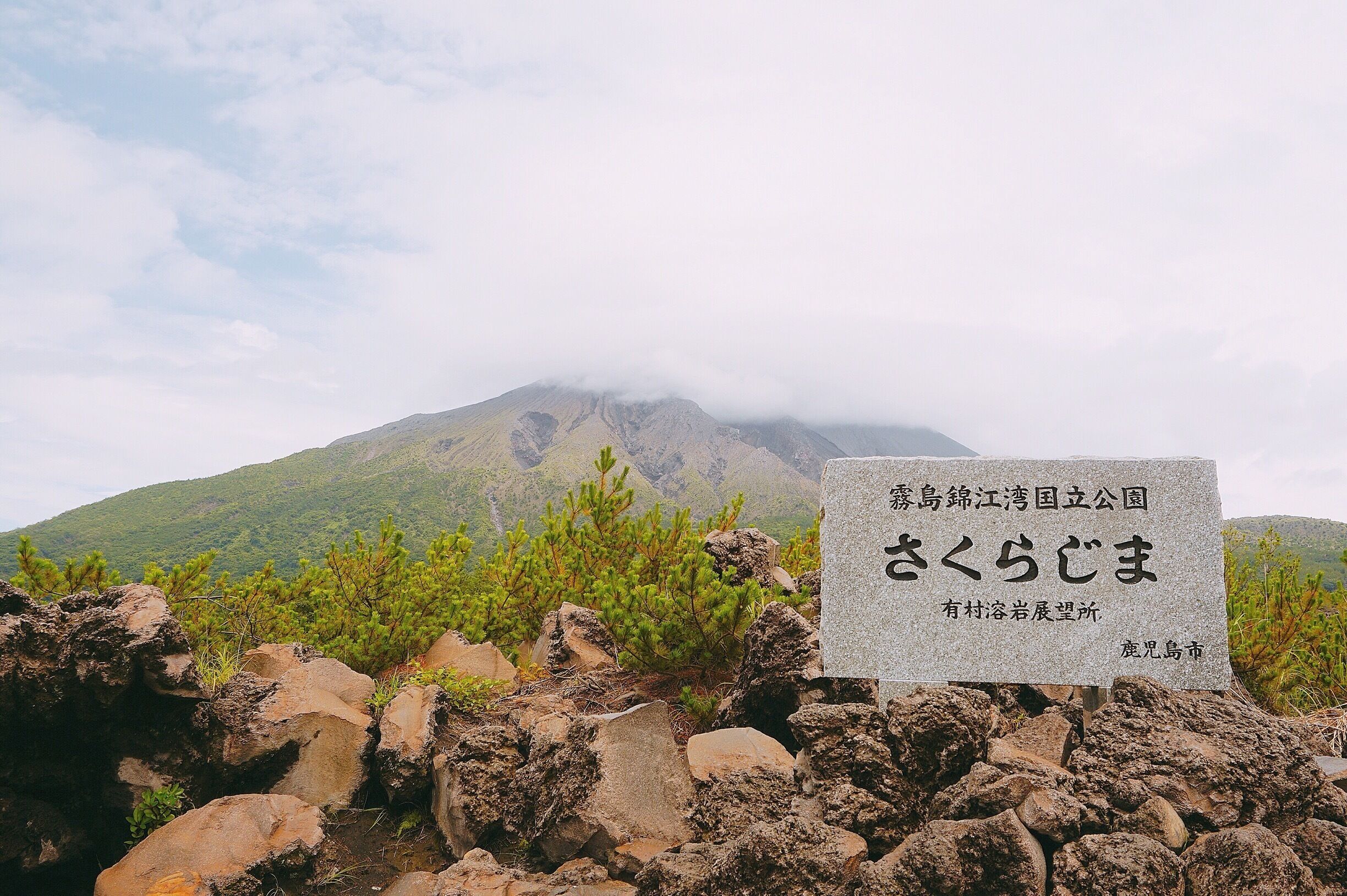 A stone sign with Japanese text in front of a mountain with green vegetation and rocks.