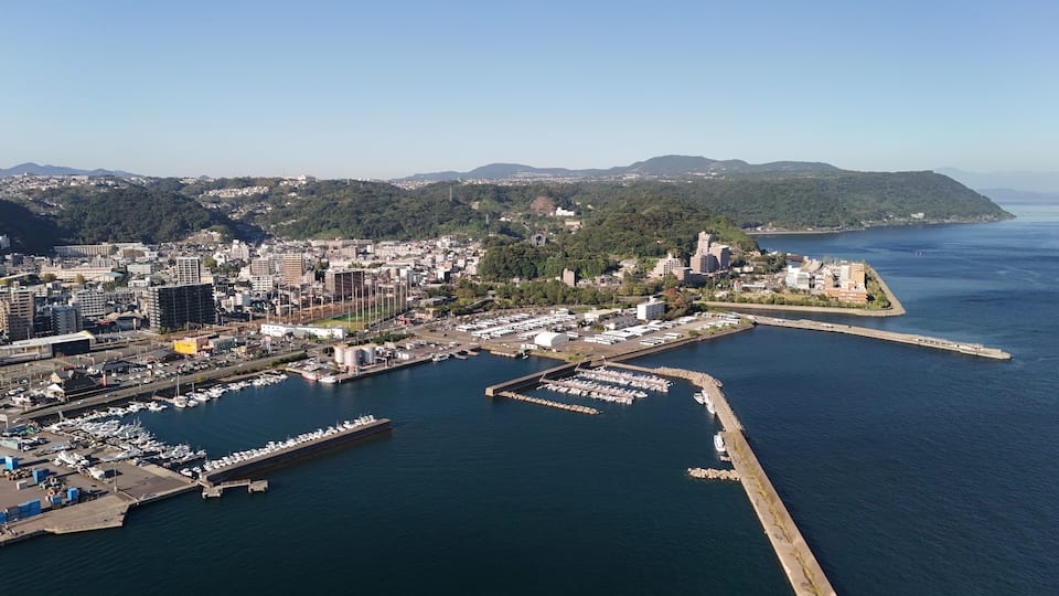 A coastal city with a marina, industrial buildings, and a hilly landscape in the background.