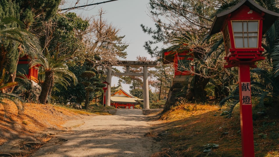 A traditional Japanese torii gate at the entrance of a shrine, surrounded by lush greenery and a dirt path leading to the main building.