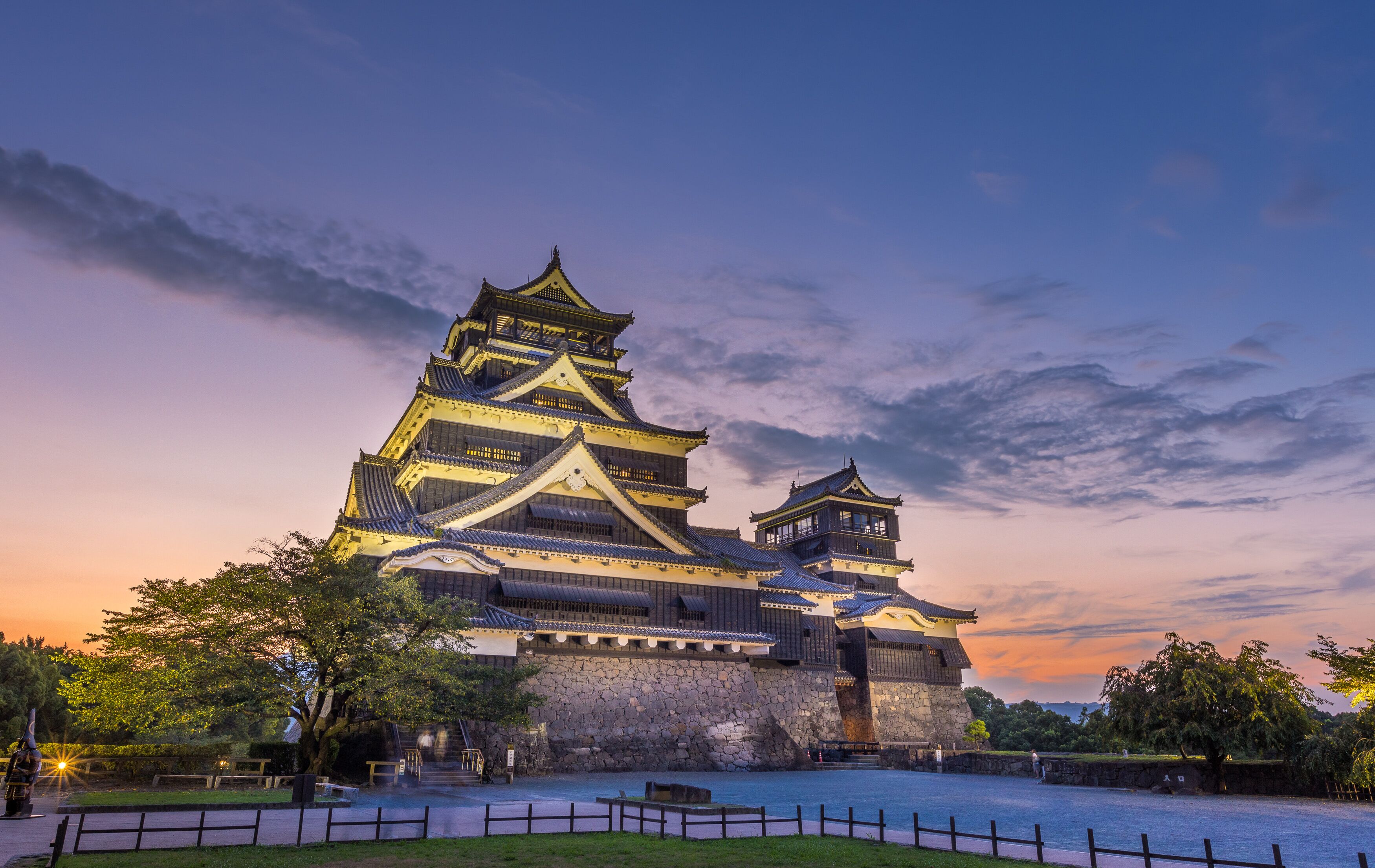 Un castillo japonés tradicional con varios niveles y base de piedra, rodeado por un foso y un parque con árboles.