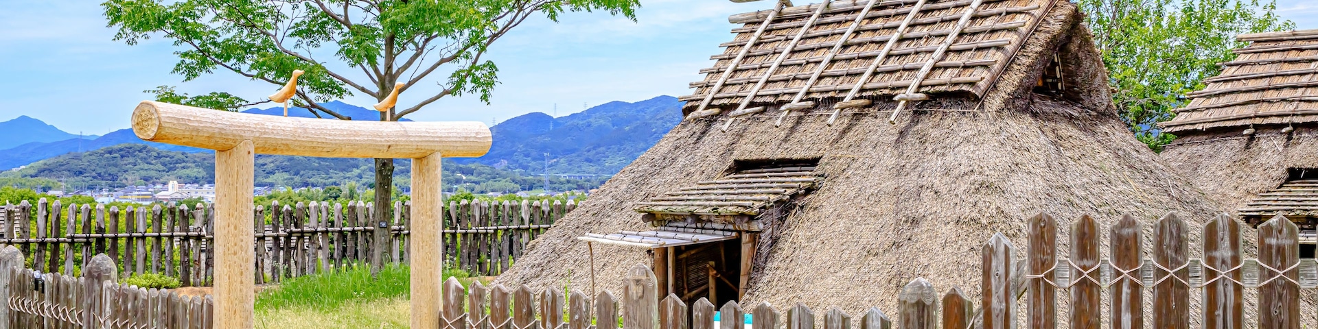 A traditional thatched-roof house with a wooden fence and a tree in the background.