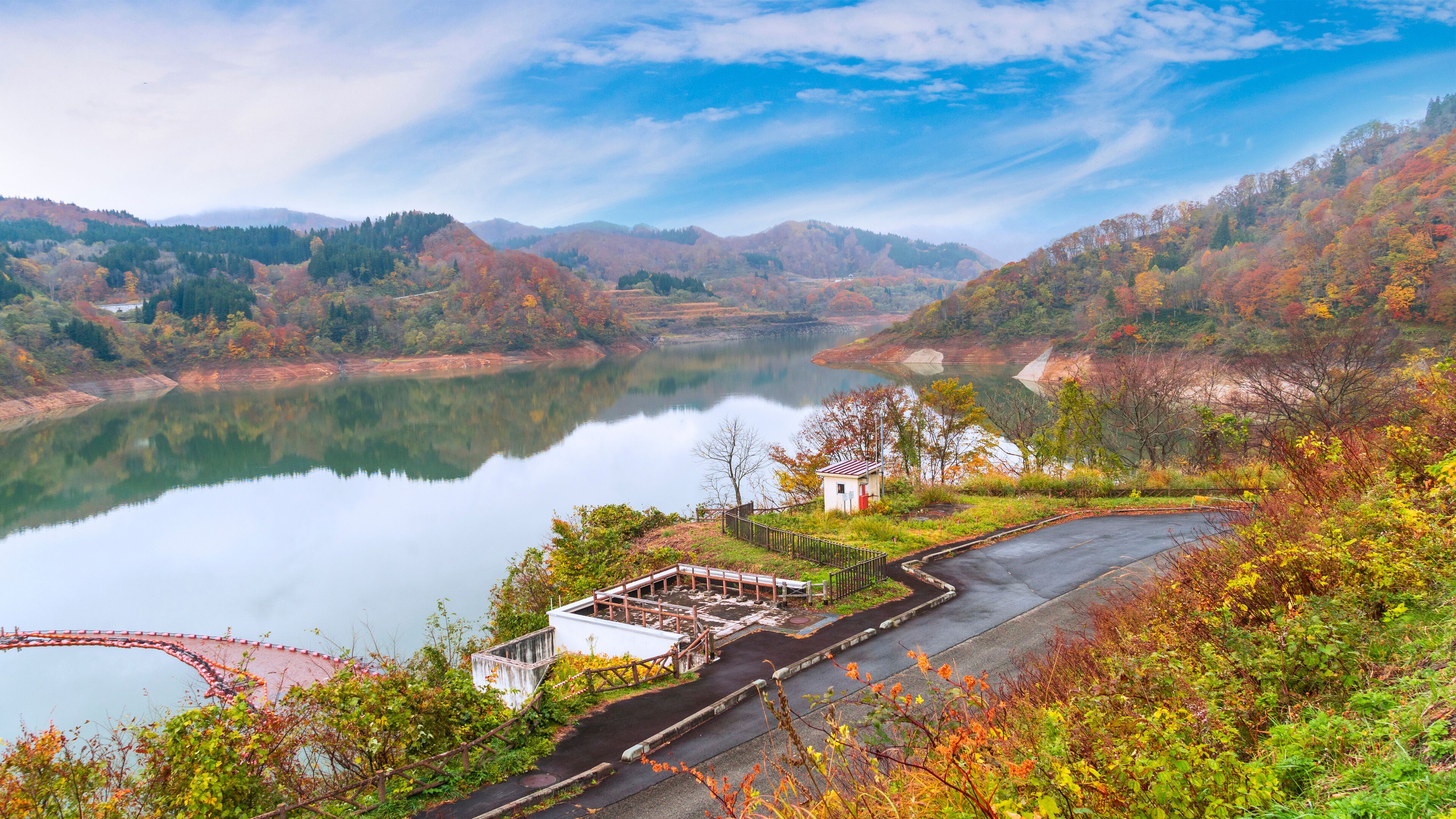 A serene lake surrounded by autumn-colored hills and a small white building near a paved path.