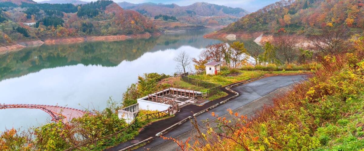 A serene lake surrounded by autumn-colored hills and a small white building near a paved path.