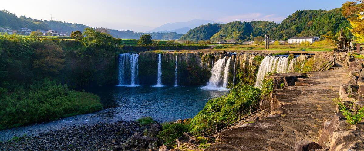 A waterfall cascading into a river surrounded by lush greenery and a stone pathway.