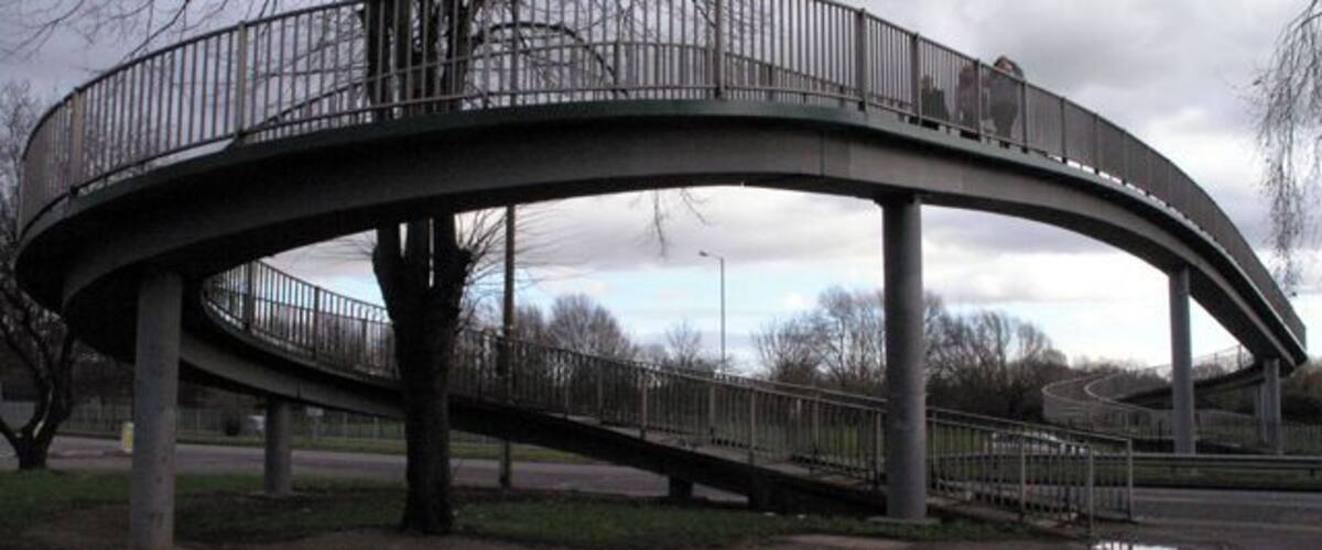 A curved pedestrian bridge with metal railings, supported by concrete pillars, in a park setting.