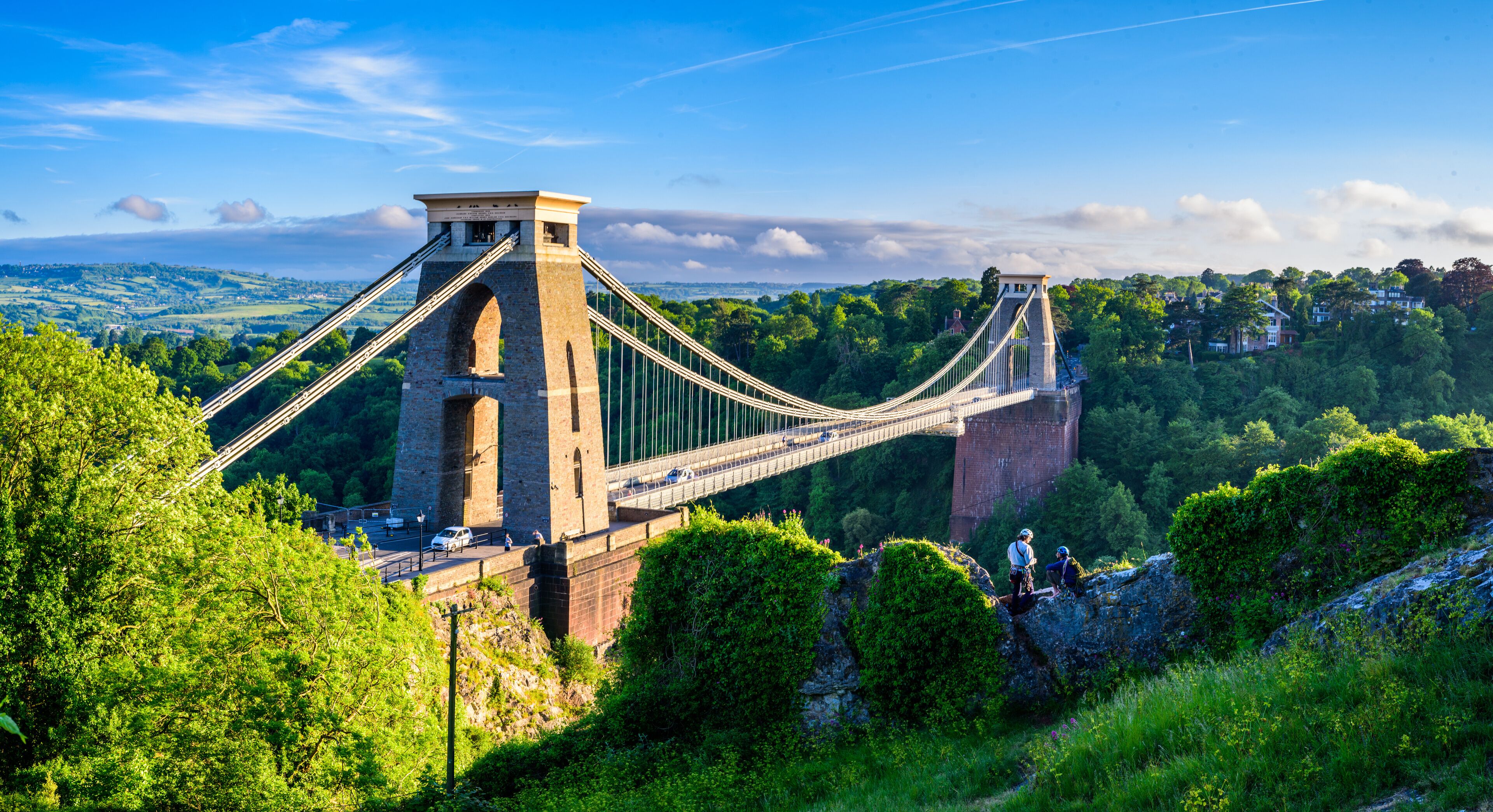 A suspension bridge with stone towers, surrounded by lush greenery and a hilly landscape.