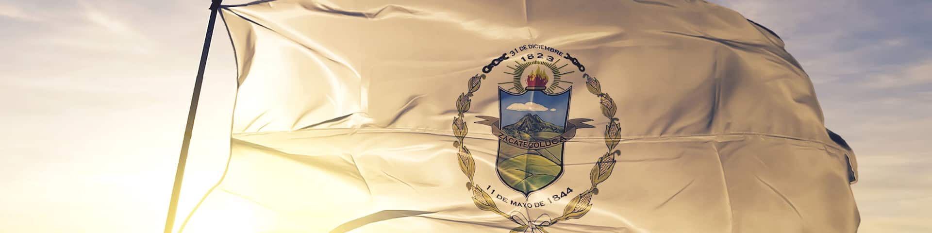 A flag with a coat of arms, a mountain, and the date "31 de diciembre de 1823" on a flagpole against a sunrise sky.