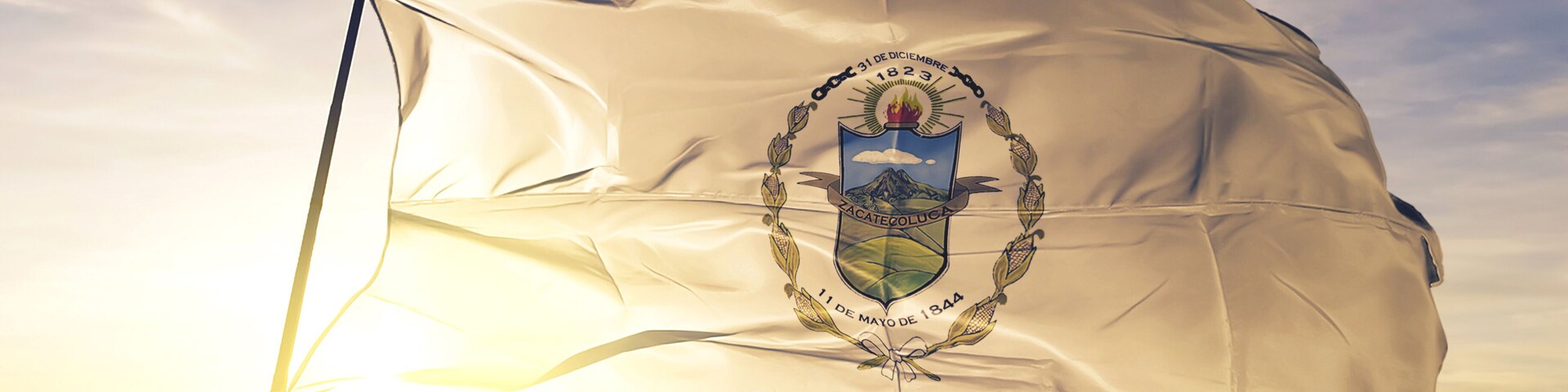Una bandera con un escudo, una montaña y la fecha "31 de diciembre de 1823" en un asta contra un cielo al amanecer.