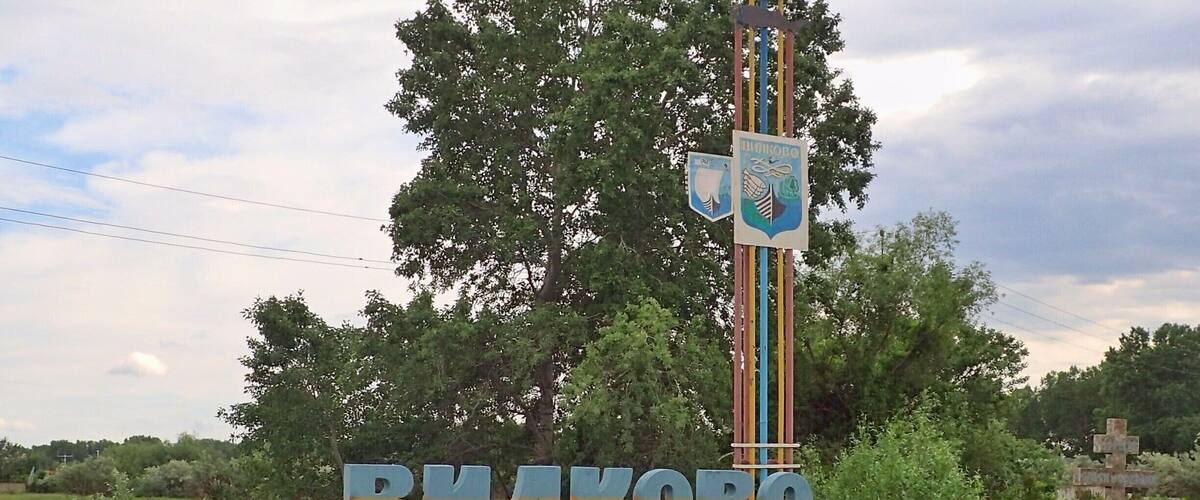 A motorcycle parked in front of a sign with 'ВЛАКОВО' and 'ОЧЕБАХ 174B' on it, surrounded by greenery.