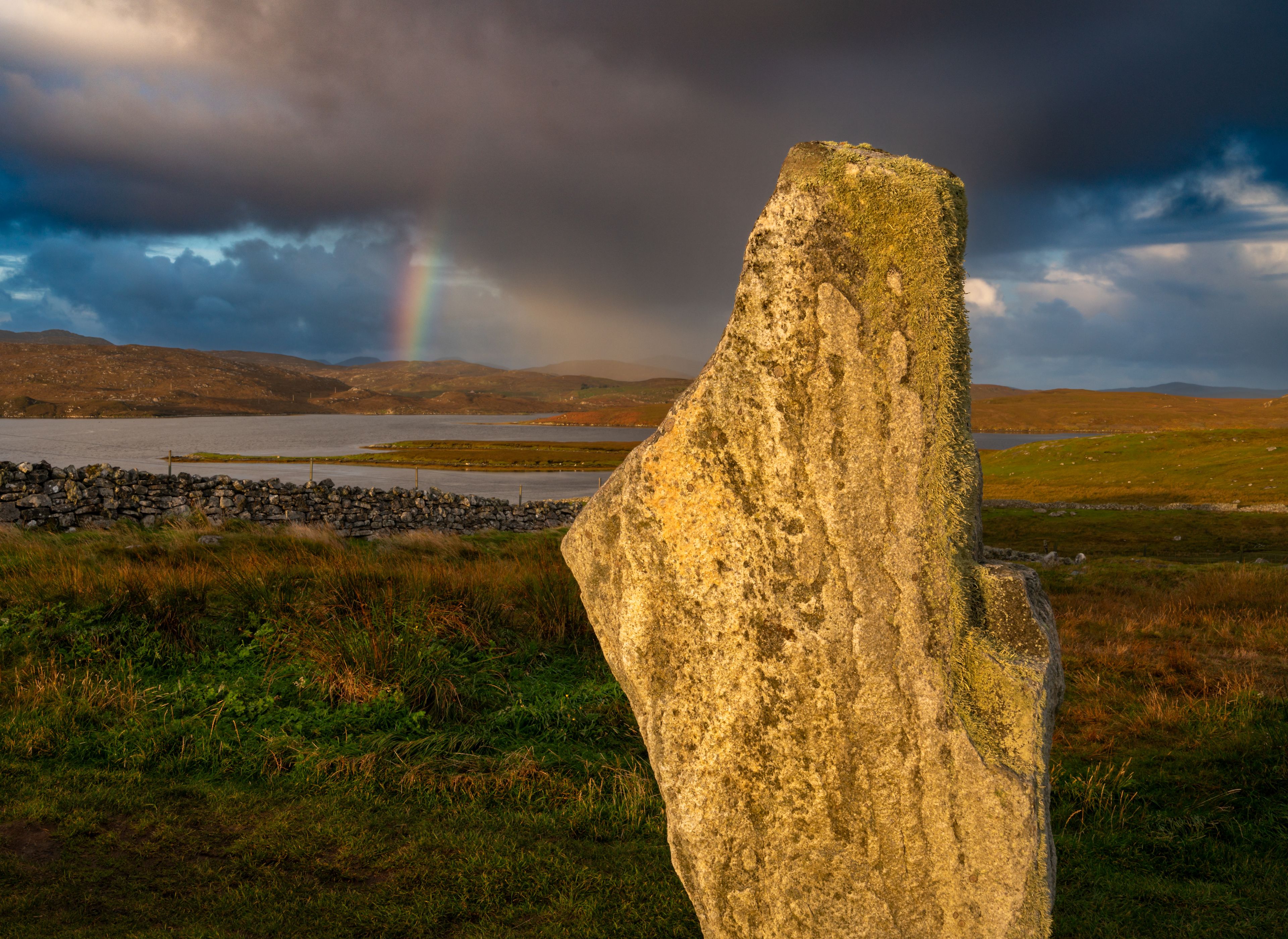 A stone monument in a grassy field with a lake and mountains in the background.