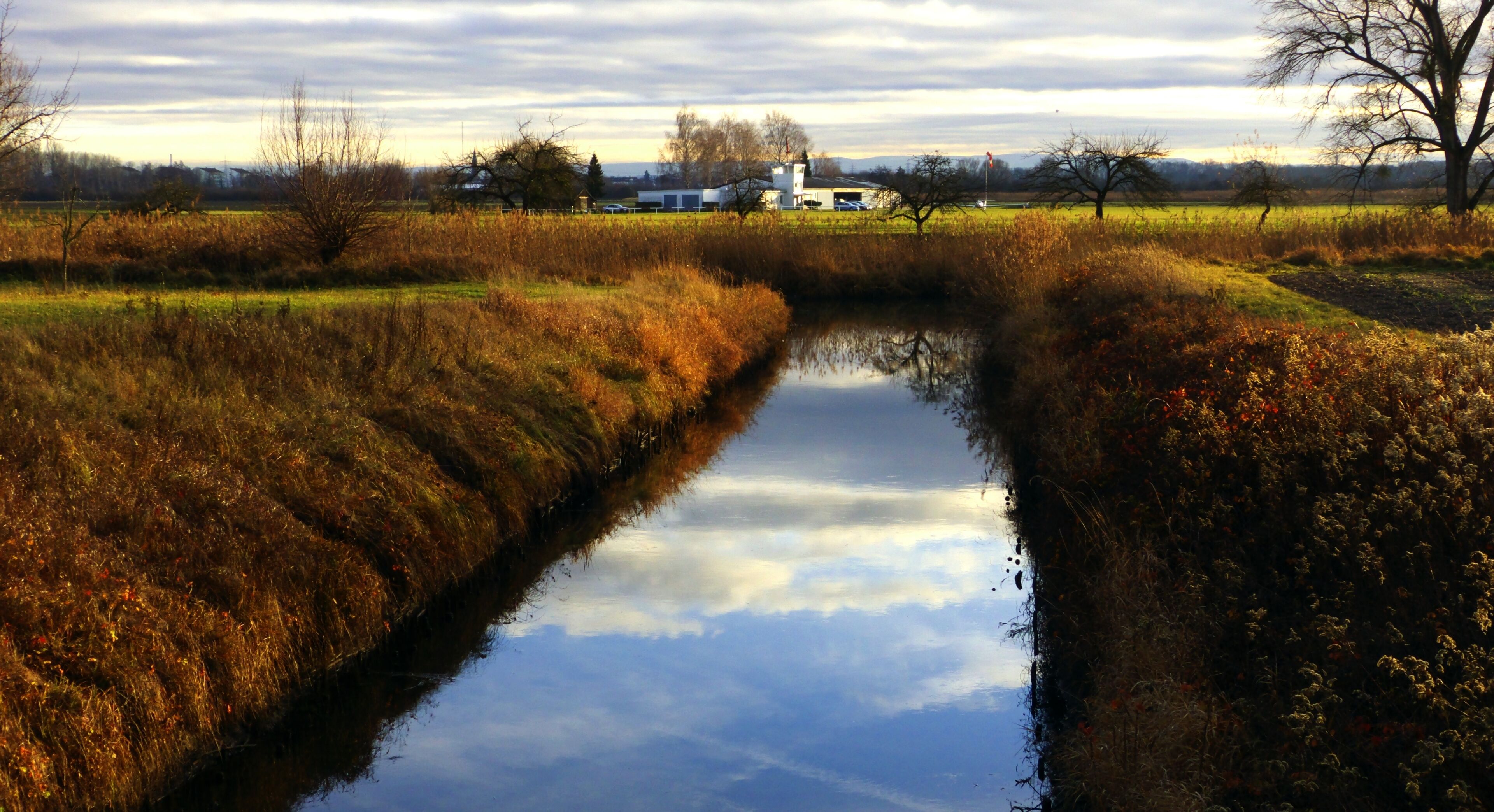 A narrow water channel with reflections of trees and sky, surrounded by grassy fields and a distant white building.