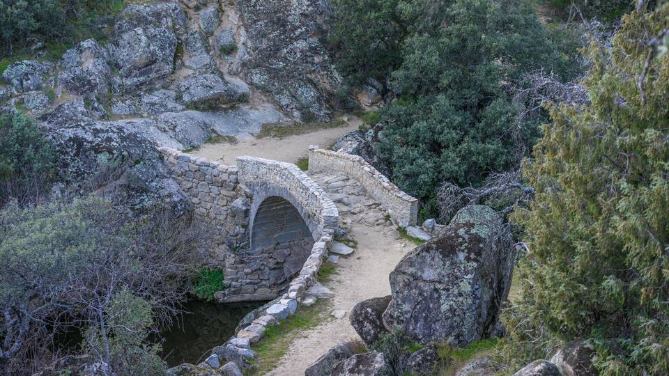 A stone bridge over a narrow stream in a rocky, forested area.