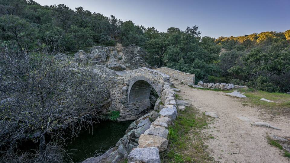 A stone bridge over a stream, surrounded by trees and rocky terrain.
