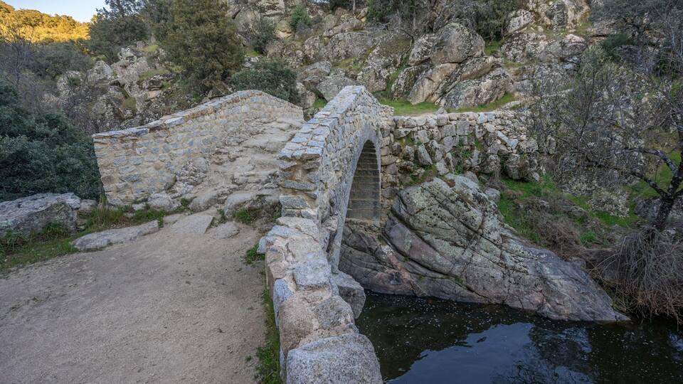 A stone bridge over a river, surrounded by rocky terrain and trees.