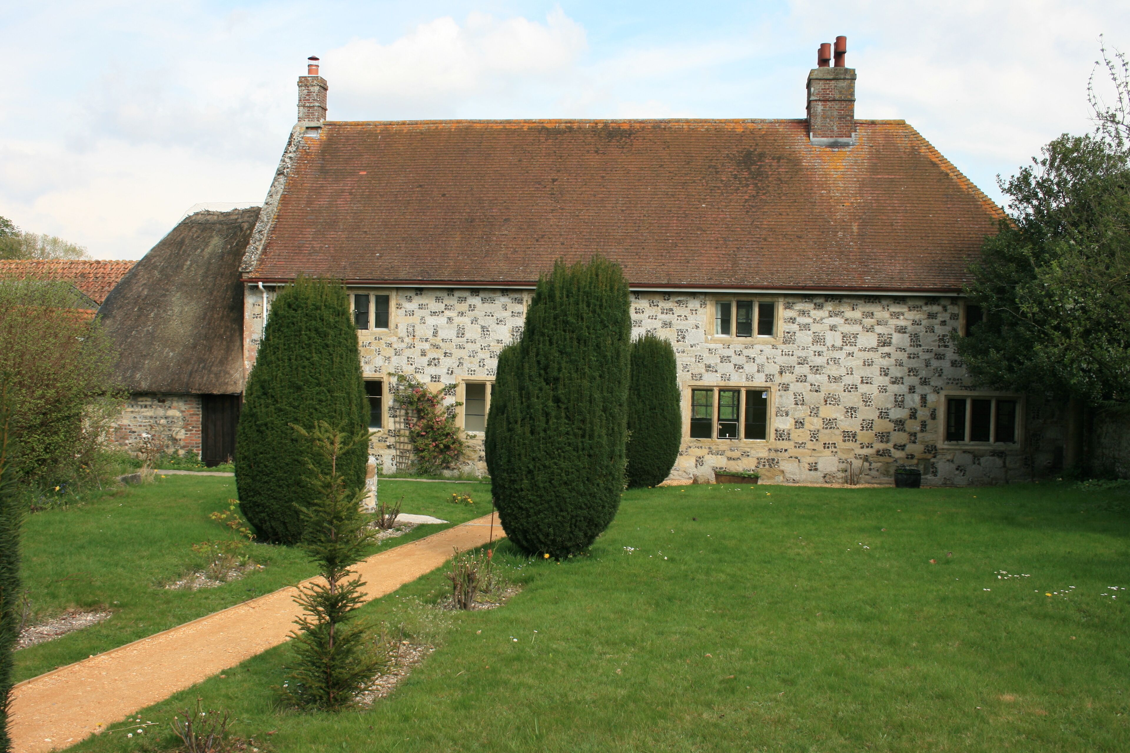 A traditional house with a tiled roof, a thatched roof structure to the left, and a well-maintained lawn with two trimmed trees.