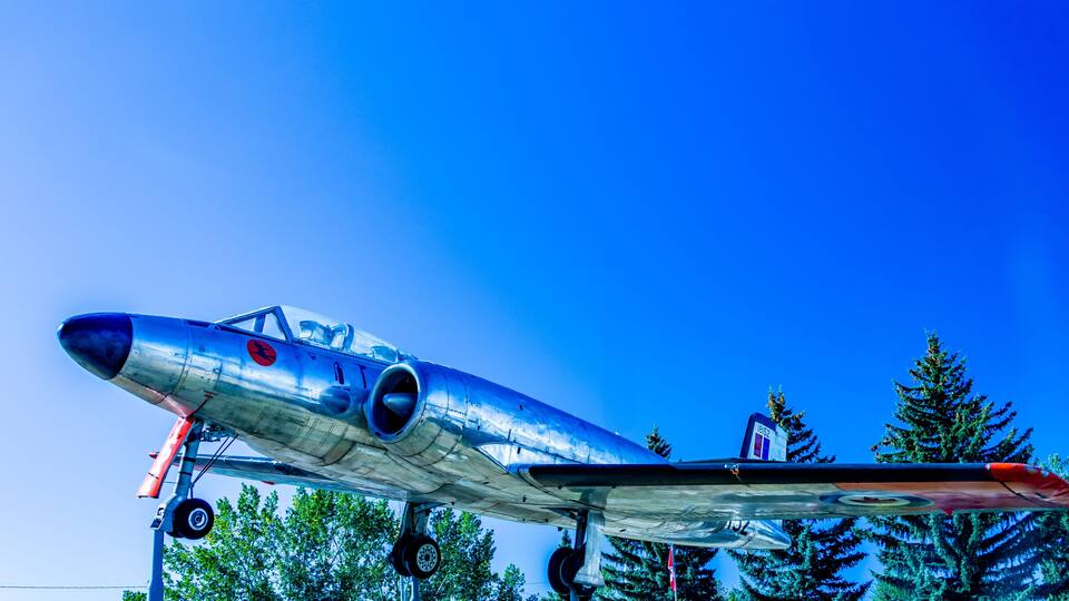 A vintage silver airplane with a red propeller, parked outdoors with trees in the background.
