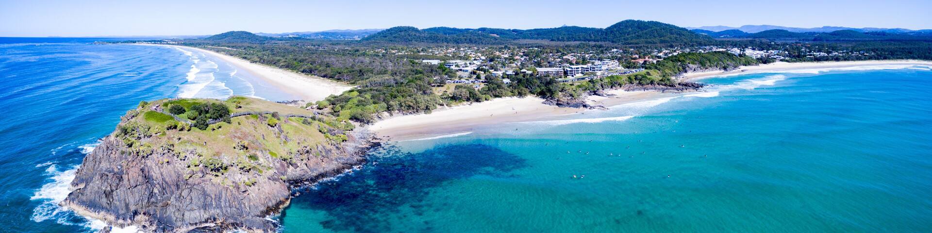 A coastal landscape with a sandy beach, clear turquoise waters, and a rocky outcrop with vegetation.
