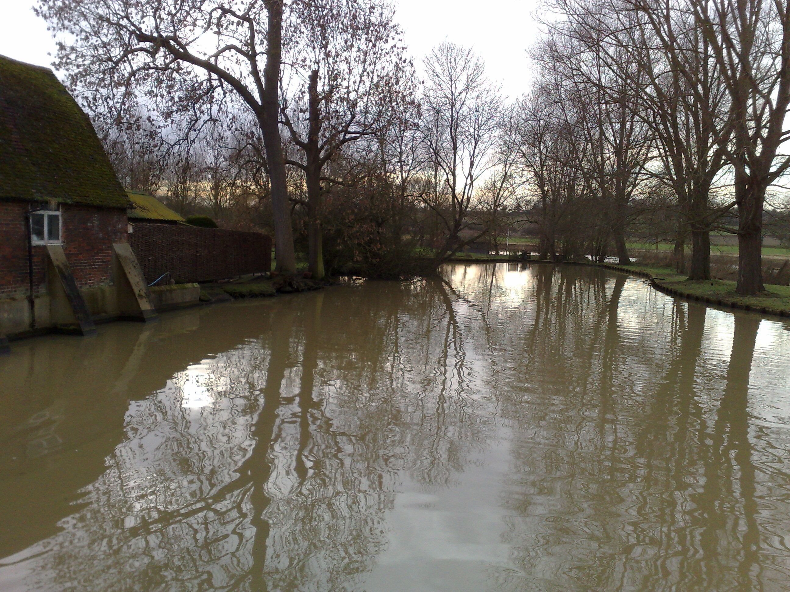 A flooded river with reflections of trees and a brick building.