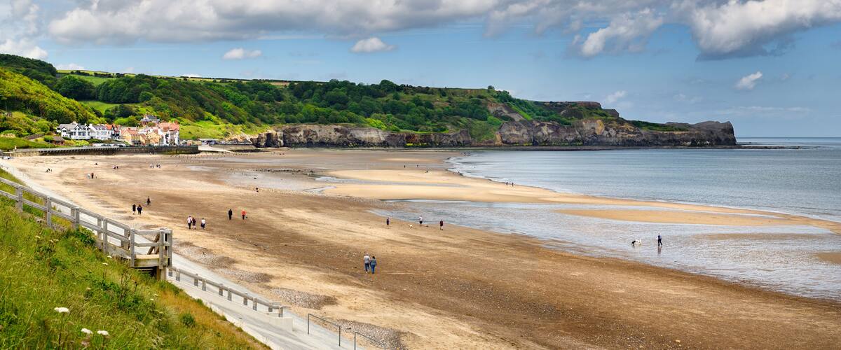 A coastal beach with people walking, a wooden walkway, and houses on a green hillside.