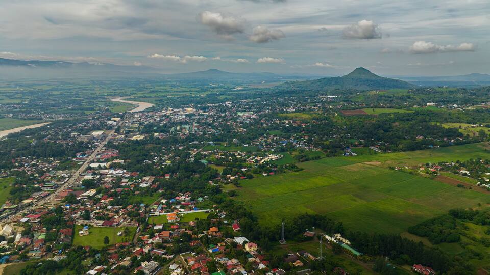 An aerial view of a town with a river, green fields, and a mountain in the background.