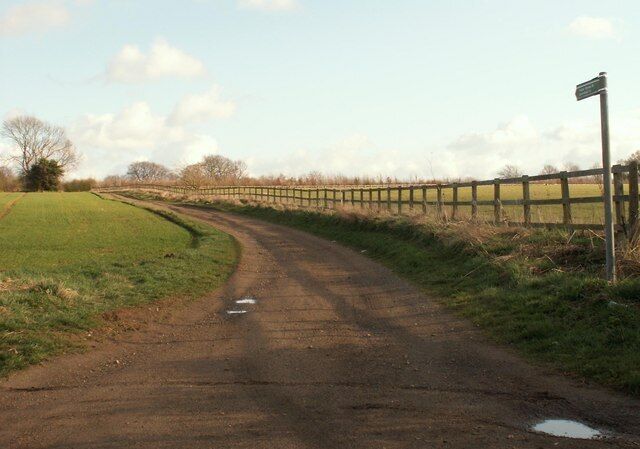 A rural dirt road curving through a grassy field with a wooden fence on one side and a signpost on the other.