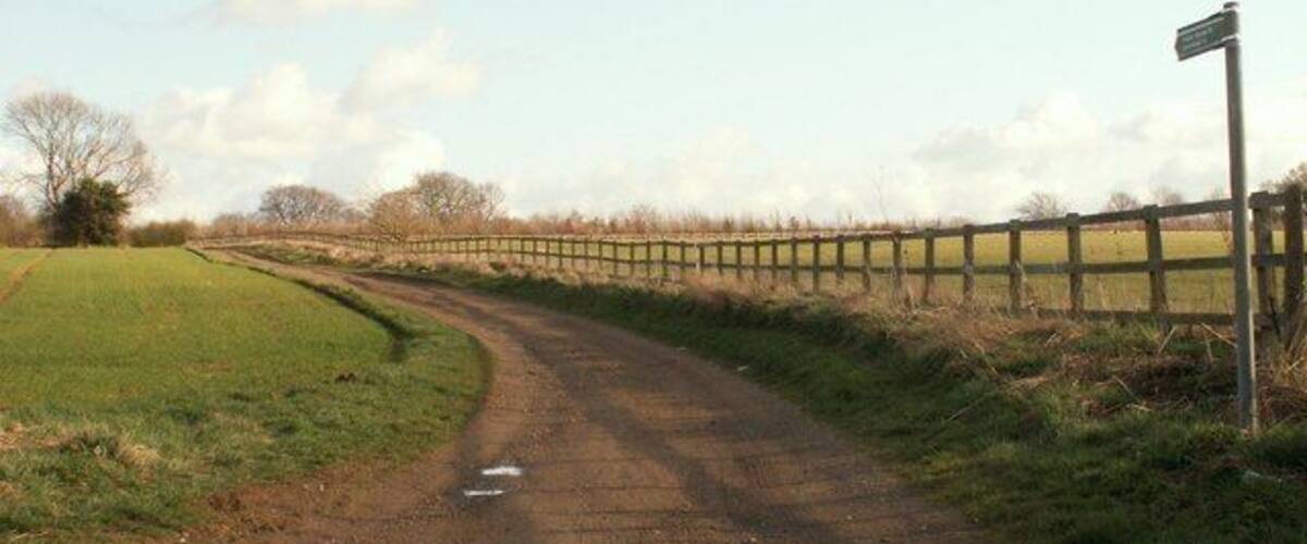A rural dirt road curving through a grassy field with a wooden fence on one side and a signpost on the other.