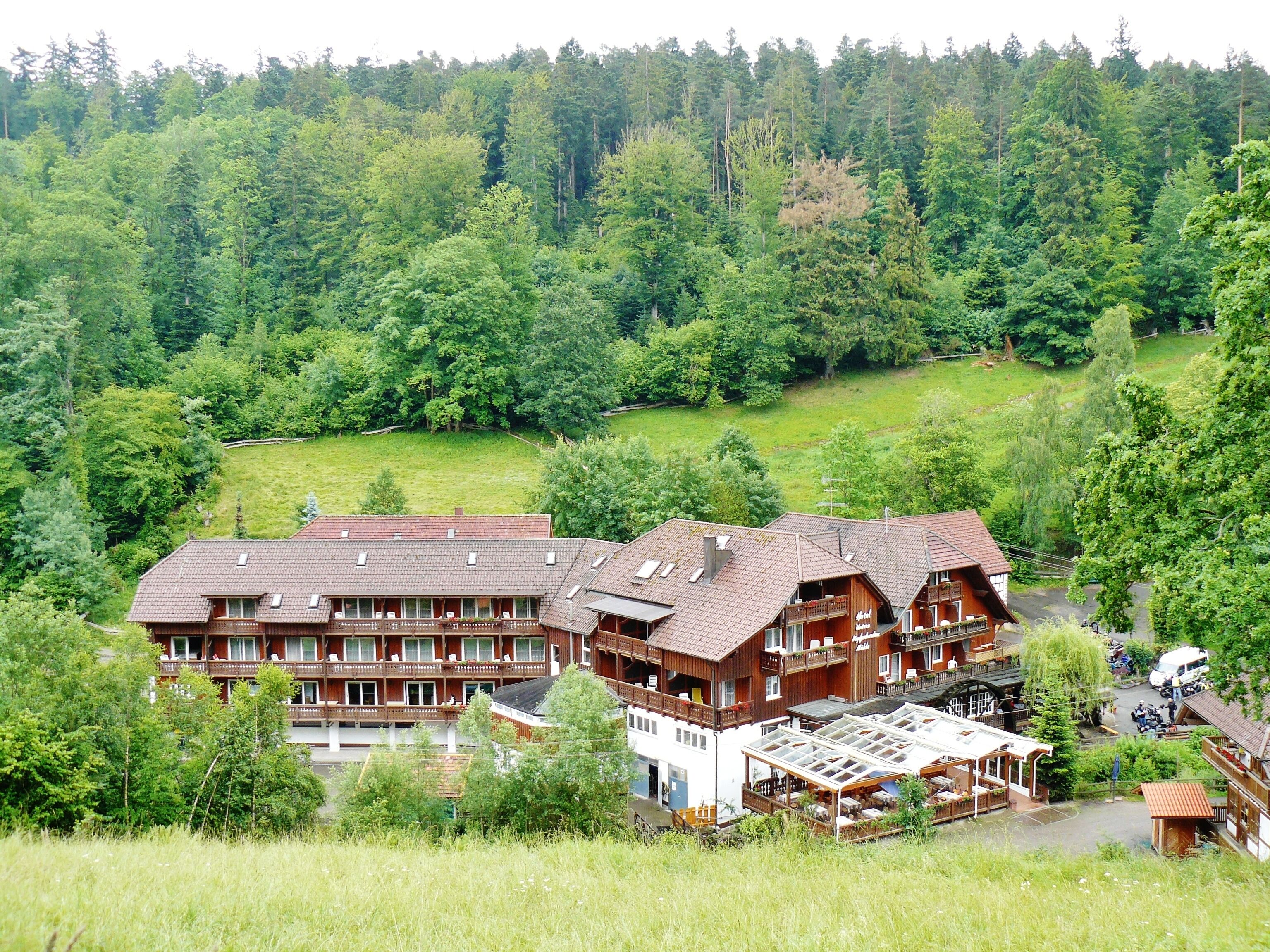 A large wooden chalet with multiple balconies, surrounded by lush greenery and a forested hillside.