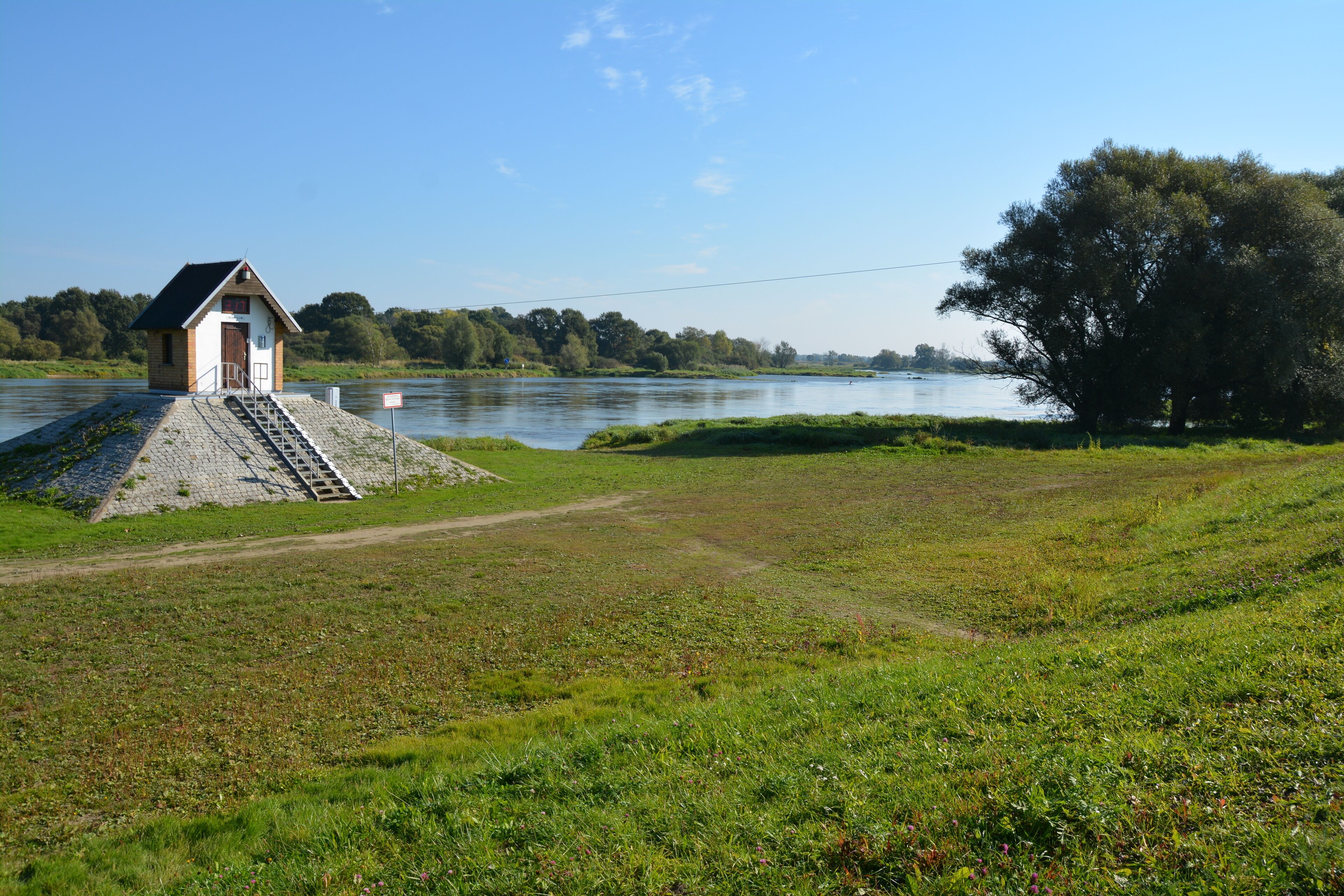 A small wooden structure by a river with a grassy field and trees in the background.
