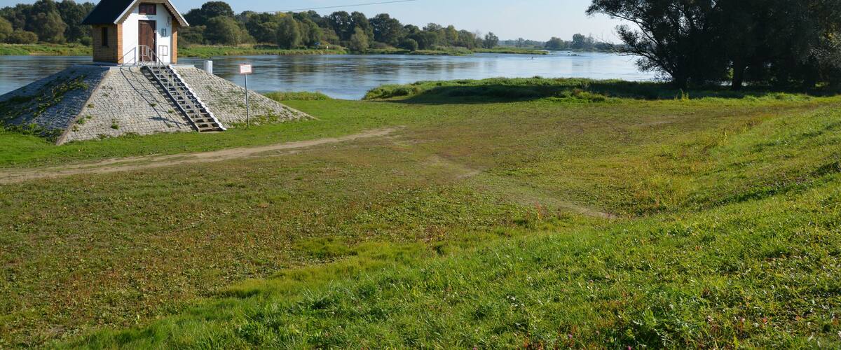 A small wooden structure by a river with a grassy field and trees in the background.