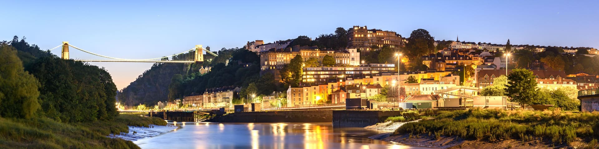 A river with a bridge, a cityscape with buildings, and a hill with trees.