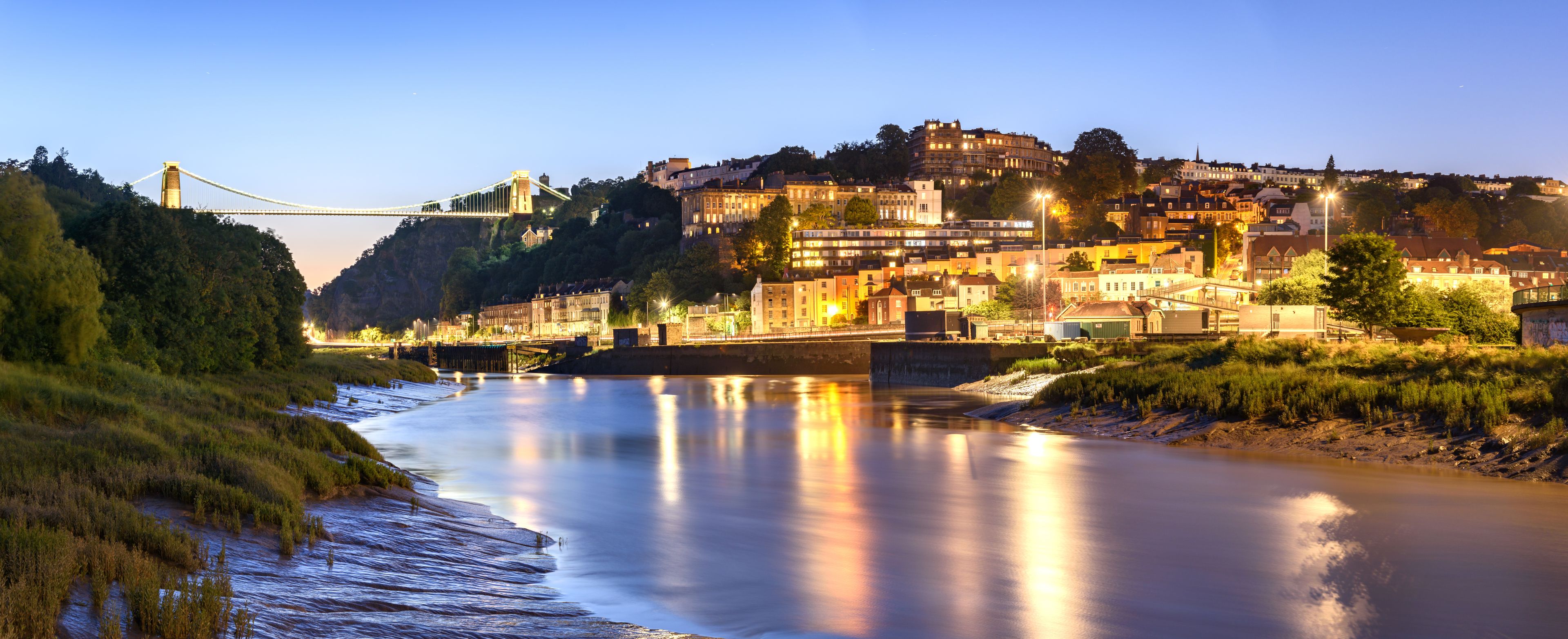 A river with a bridge, a cityscape with buildings, and a hill with trees.