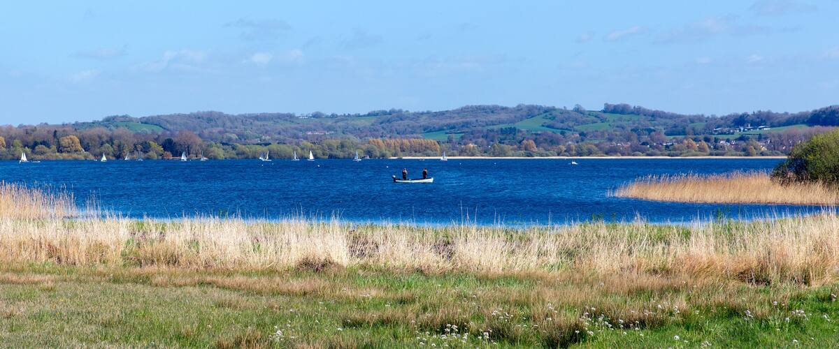 A lake with sailboats, grassy banks, and a hilly landscape in the background.