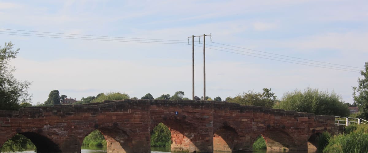 A brick bridge over a calm river with reflections, surrounded by greenery and power lines.